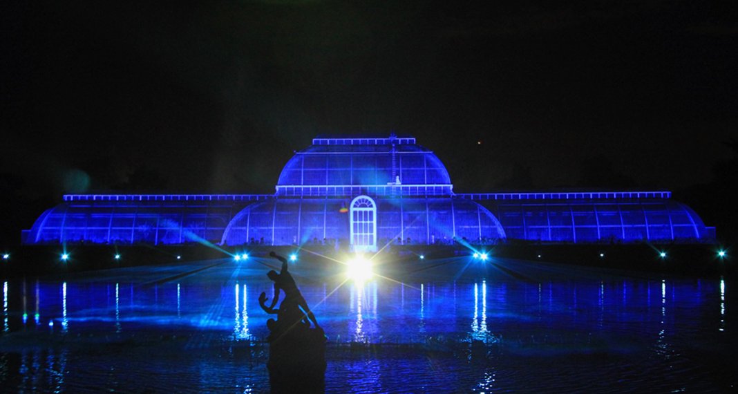 Kew Gardens illuminated at night with blue lighting, featuring a large glass conservatory and a decorative statue in the foreground, highlighting Midas' role in providing eco-friendly power solutions for the Christmas at Kew event.