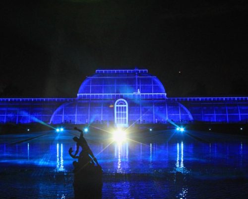 Kew Gardens illuminated at night with blue lighting, featuring a prominent glasshouse and a water reflection, showcasing Midas' innovative power solutions.