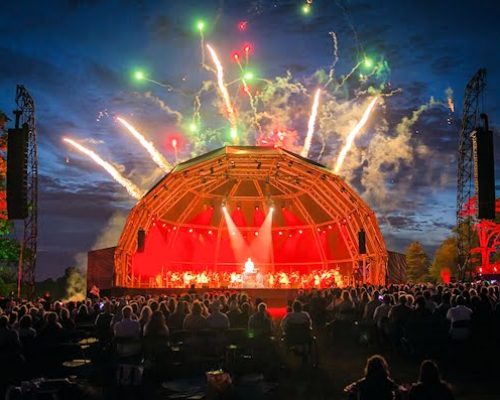 Outdoor concert at Leeds Castle featuring a large stage with vibrant lighting and fireworks, attended by a crowd, showcasing Midas's power solutions for events.