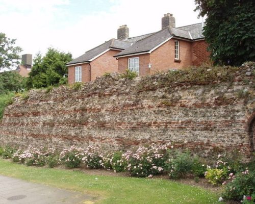 Colchester Roman Wall with flowering shrubs and residential buildings in the background, illustrating Midas Generator Hire's involvement in heritage projects.