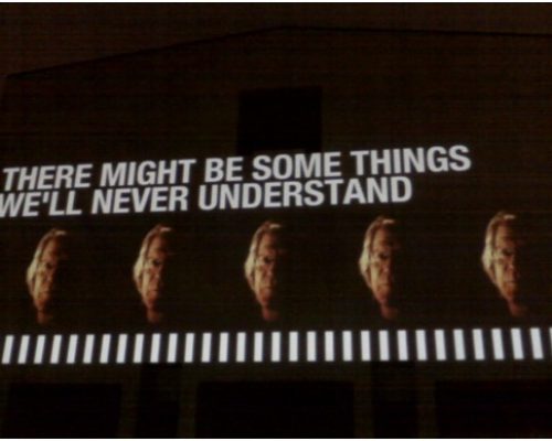 Public artwork display featuring projected text "There might be some things we'll never understand" alongside four faces, emphasizing contemporary themes of heroism and public art engagement.