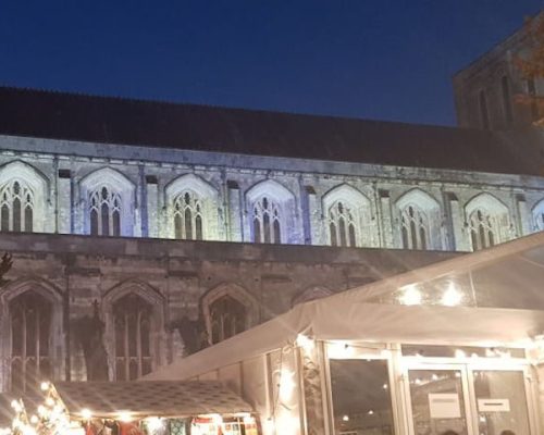 Historic building illuminated at night, featuring gothic architecture and decorative lighting, with a festive market stall in the foreground, showcasing Midas’ contribution to event lighting.