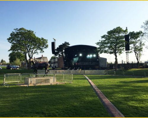 Outdoor event stage setup with sound equipment, surrounded by trees and a statue, showcasing Midas's role in providing temporary power solutions for live events.