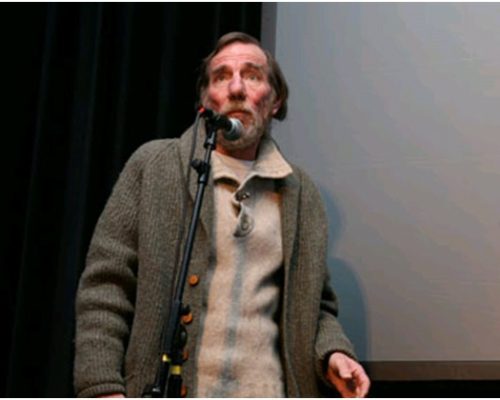 Man speaking into a microphone during a presentation, wearing a gray sweater, against a dark backdrop, related to the film "The Age of Stupid."
